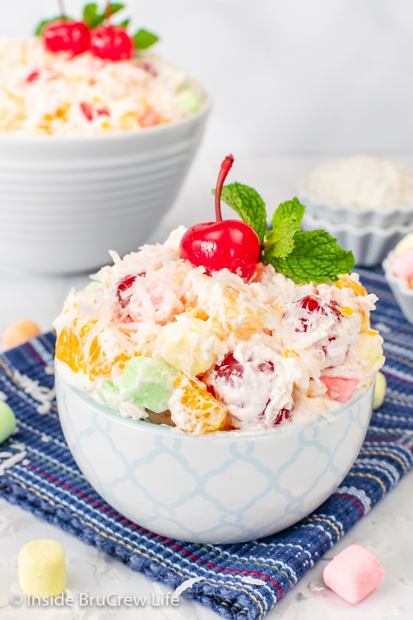 A blue and white bowl on a blue towel filled with ambrosia fruit salad and topped with a cherry and mint leaves.