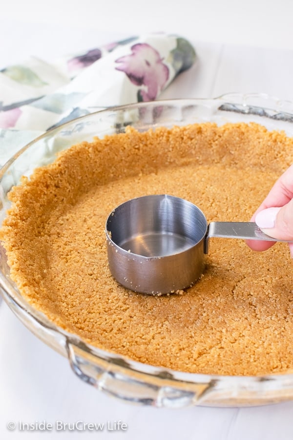 A glass pie plate with a graham cracker pie crust in it and a measuring cup pressing it down.