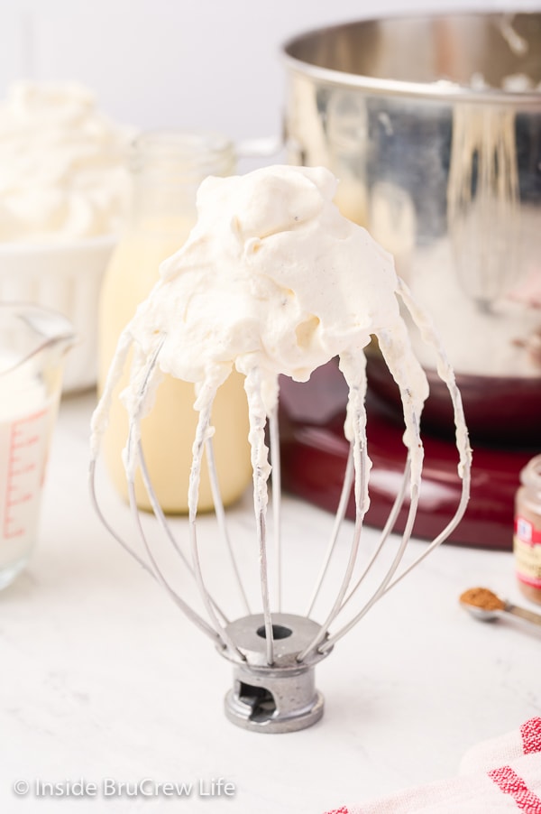 A wire whisk attachment standing on a white board with fluffy eggnog whipped cream on it.