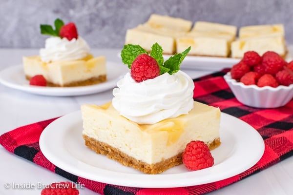 Two white plates with squares of lemon cheesecake on them topped with whipped cream and raspberries.
