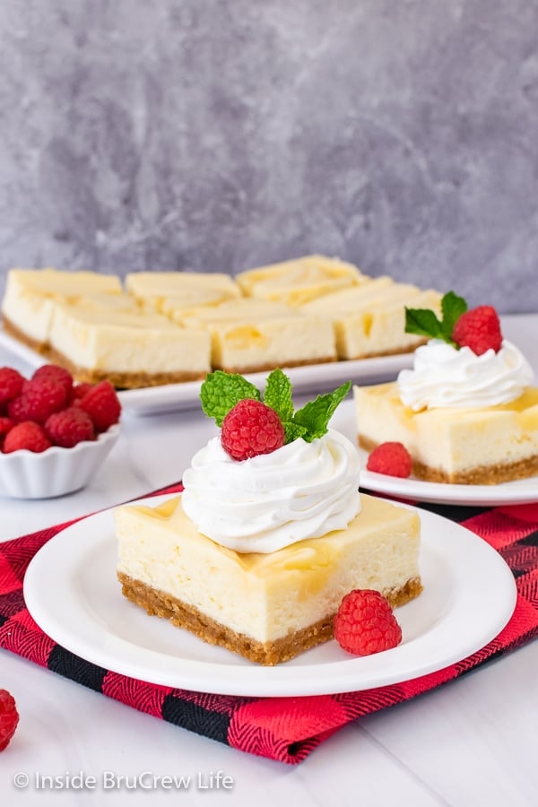 Two white plates with lemon cheesecake topped with whipped cream and raspberries on it.