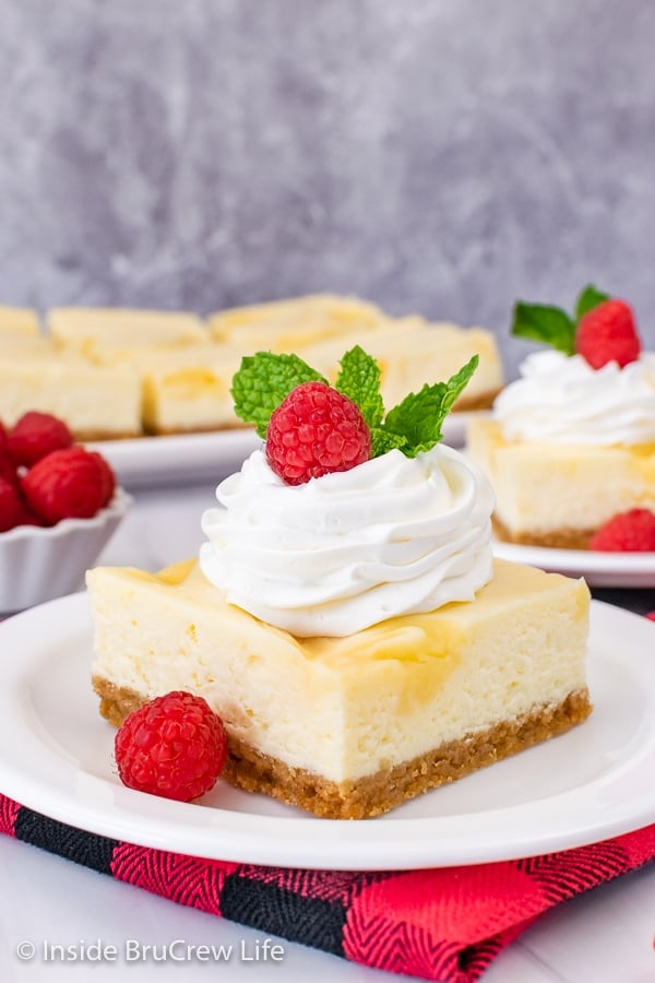A white plate with a lemon cheesecake bar topped with whipped cream and a raspberry on it.