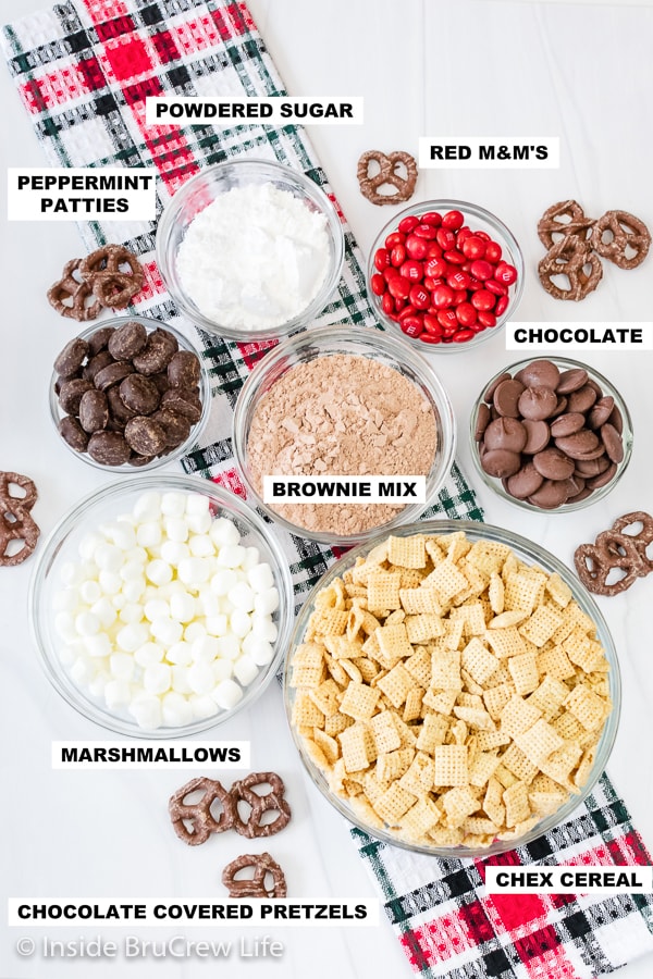 An overhead picture of a white board with bowls of ingredients needed to make a Christmas Chex mix.