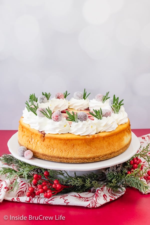 A white cake plate with a full sized cranberry cheesecake topped with whipped cream, candied cranberries, and thyme sprigs.