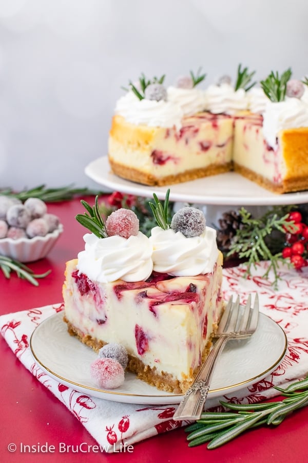 A slice of cranberry cheesecake on a white plate topped with whipped cream, sugared cranberries, and thyme.