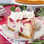 A white plate with a slice of cranberry cheesecake topped with cool whip and candied cranberries on it.