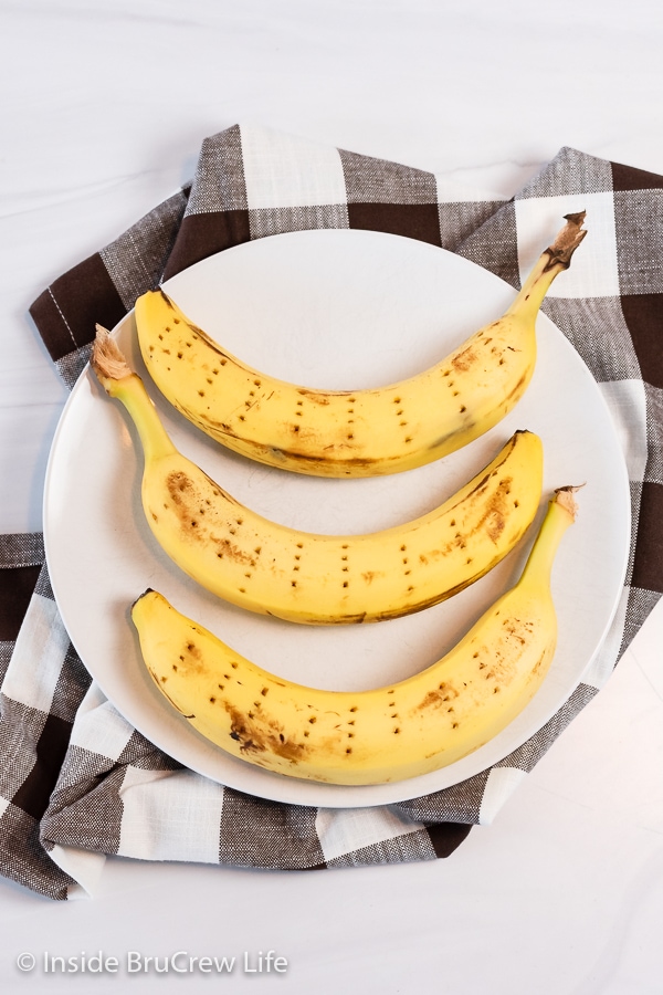 A round white plate with three yellow bananas on it with fork holes all over them.