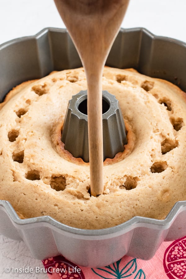 A cake in a bundt pan getting holes poked all around the center with a wooden spoon handle.