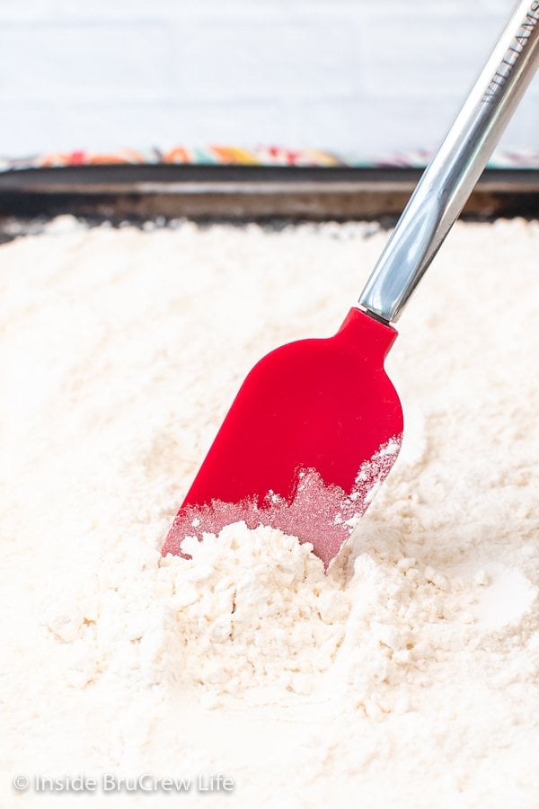 A spatula moving flour around on a sheet pan.