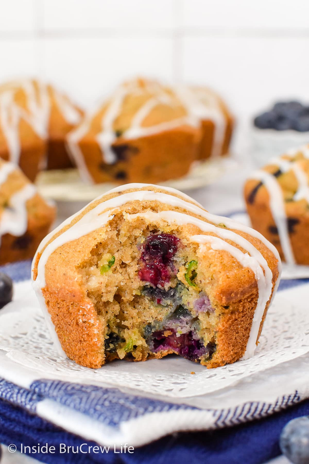 A zucchini muffin with blueberries on a white paper with a bite out of it.