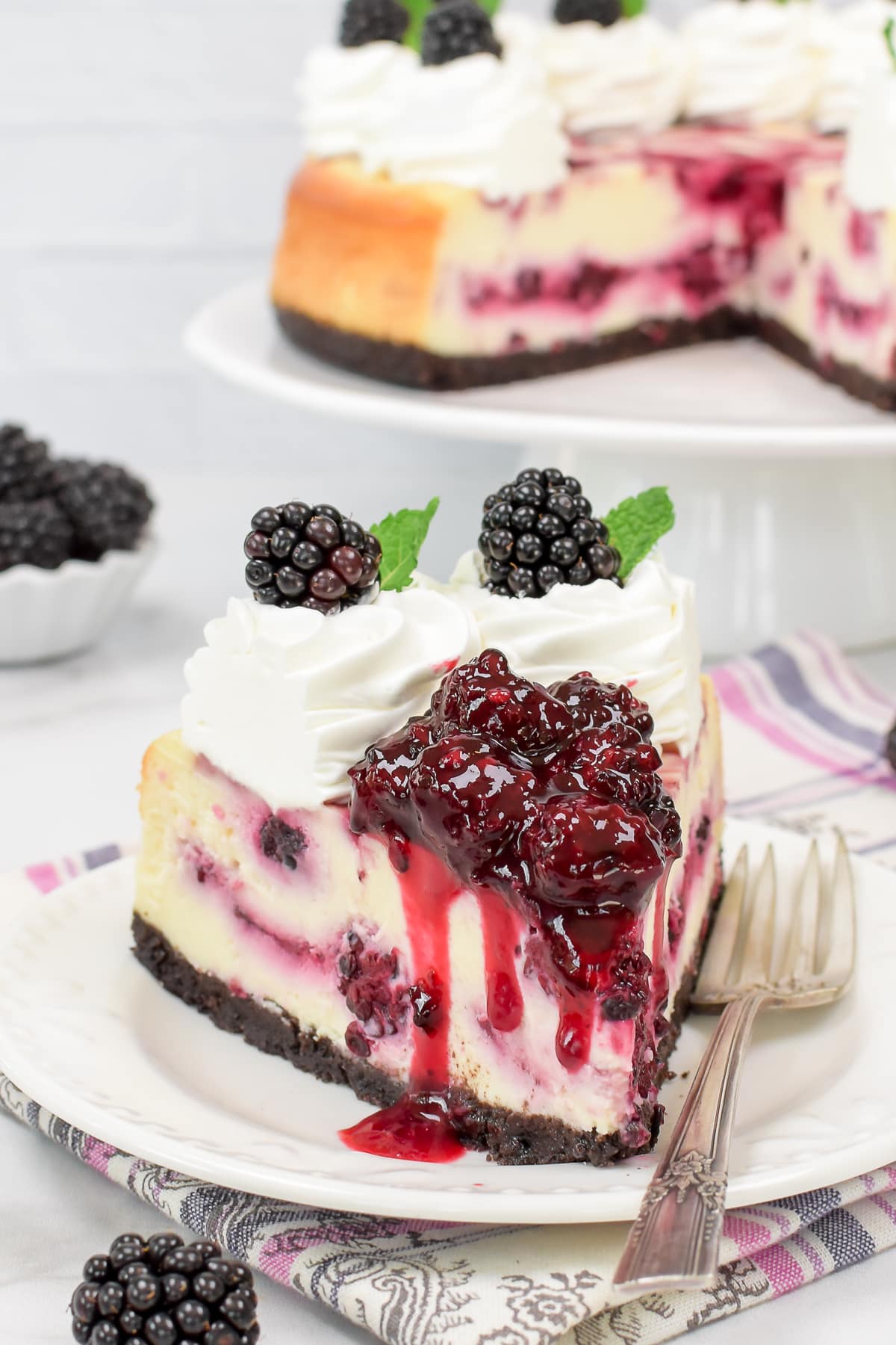 A white plate with a slice of cheesecake topped with blackberry pie filling on top.