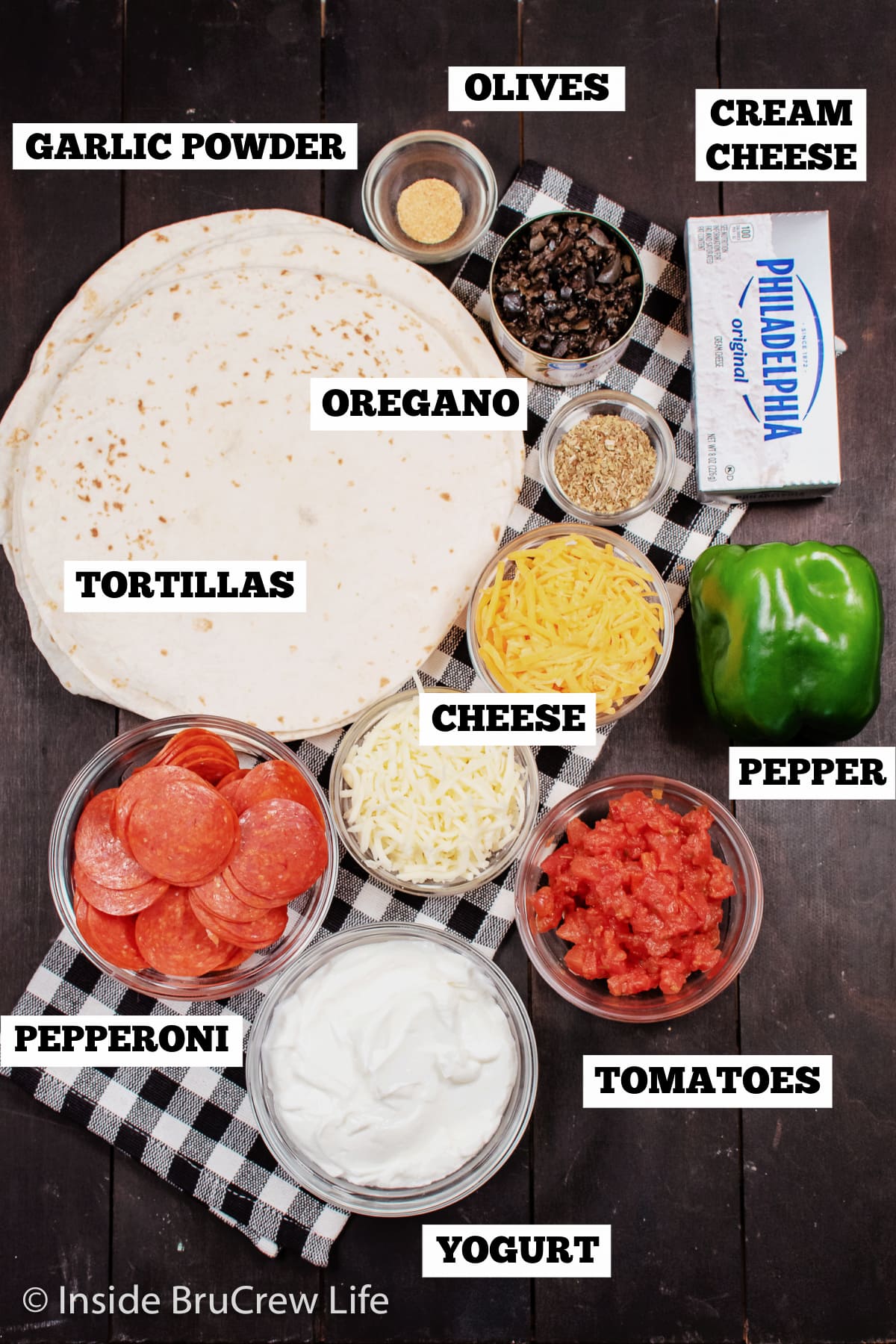 A brown board with bowls of ingredients needed to make roll ups with tortillas and pizza toppings.