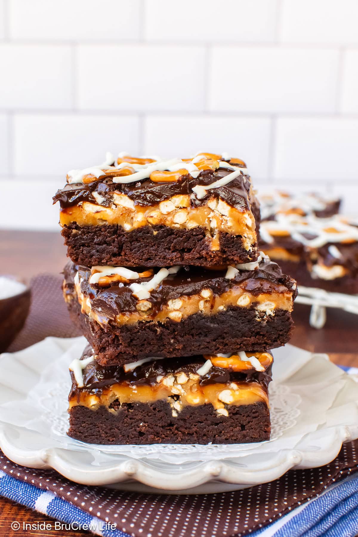 Three chocolate brownies topped with caramel and pretzels stacked on a plate.