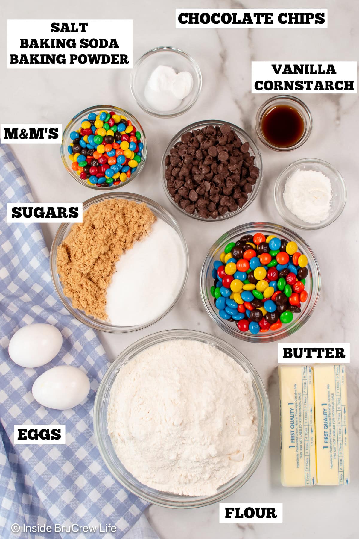 Bowls of ingredients needed to make chocolate chip cookies with M&M's on a white board.