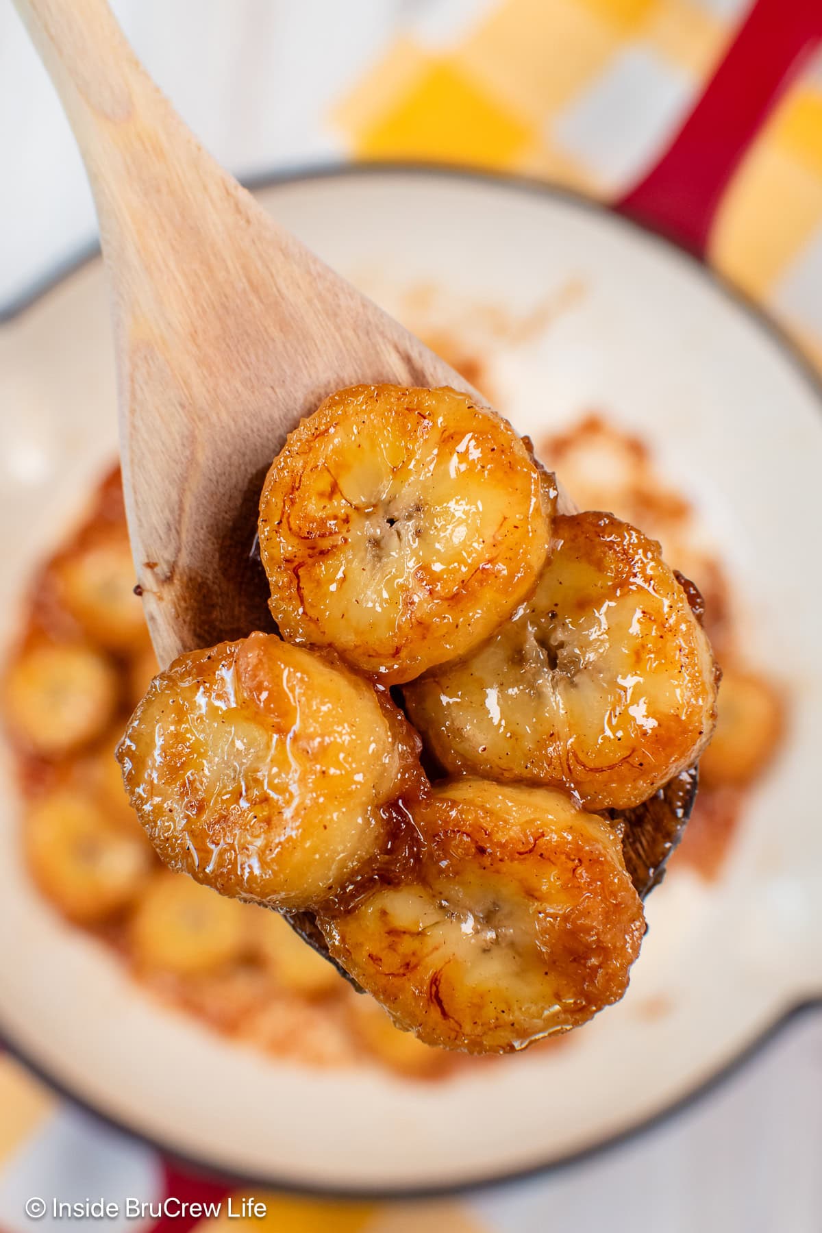 A wooden spoon lifting caramel banana slices out of a pan.