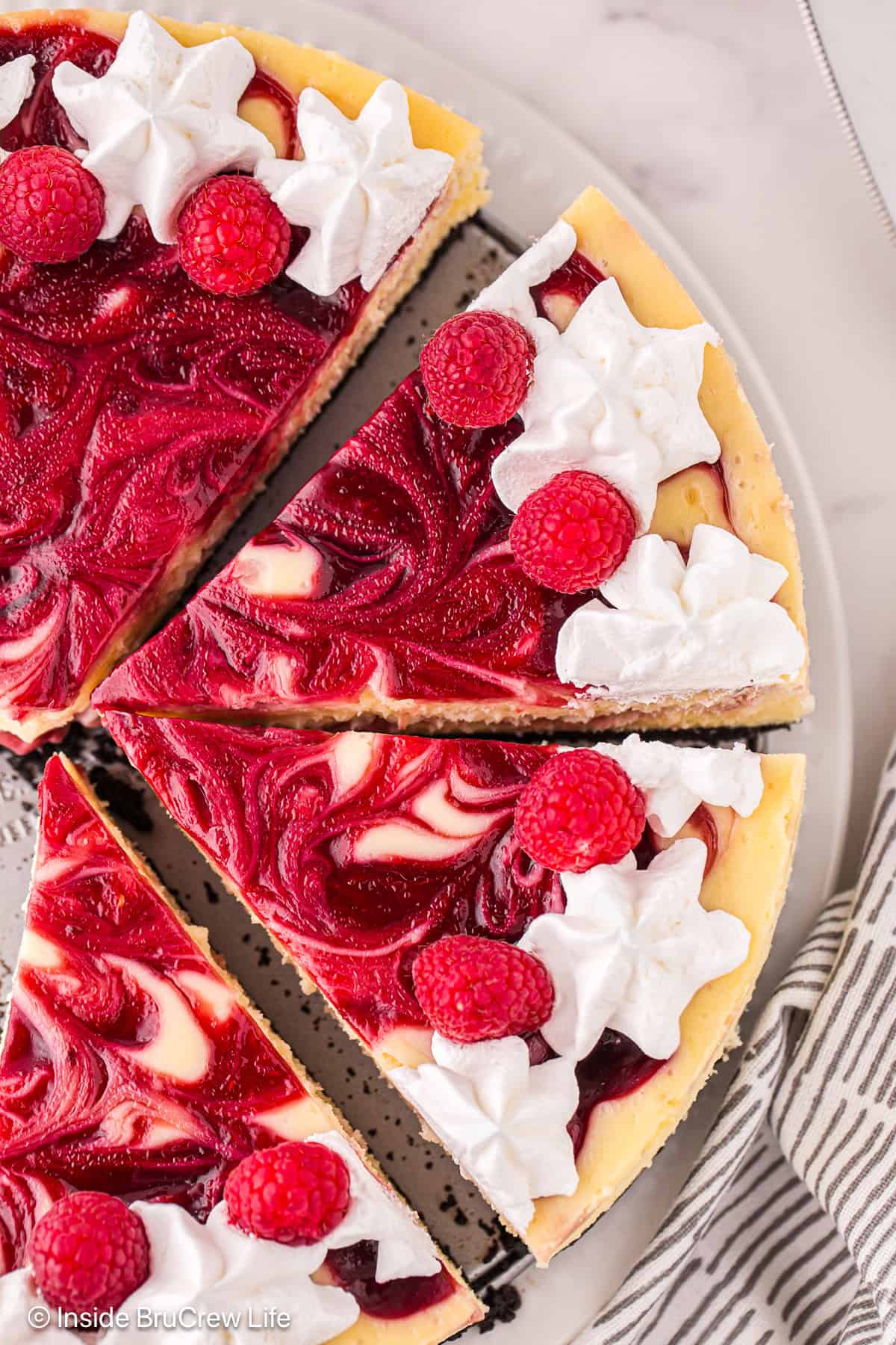 Close up overhead view of a white chocolate raspberry cheesecake topped with a border of piped whipped cream rosettes, cut into slices.