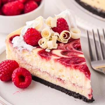 A slice of white chocolate raspberry cheesecake topped with whipped cream, fresh raspberries, and white chocolate curls on a plate with a fork.