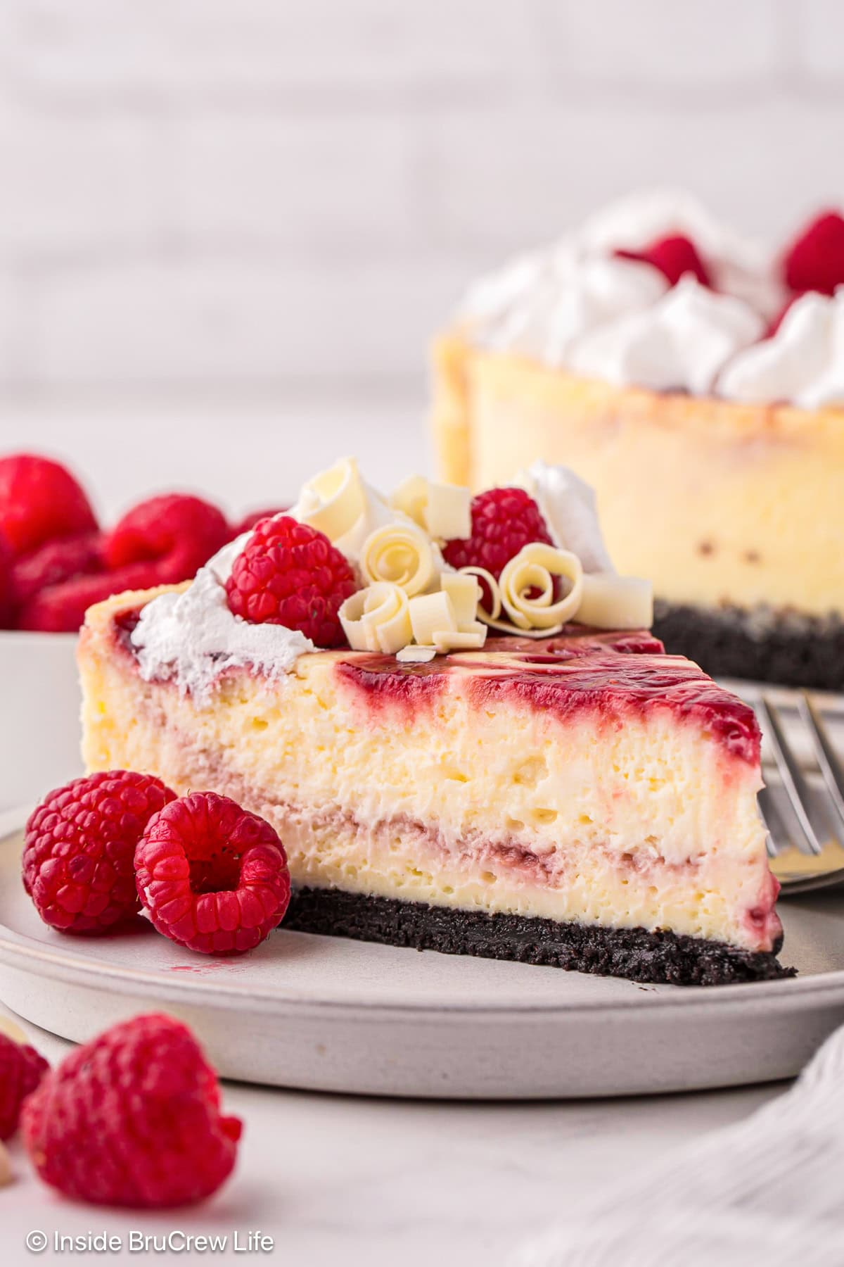 A slice of white chocolate raspberry cheesecake topped with whipped cream, fresh raspberries, and white chocolate curls on a plate with a fork.