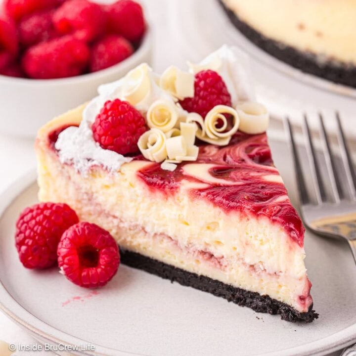 A slice of white chocolate raspberry cheesecake topped with whipped cream, fresh raspberries, and white chocolate curls on a plate with a fork.