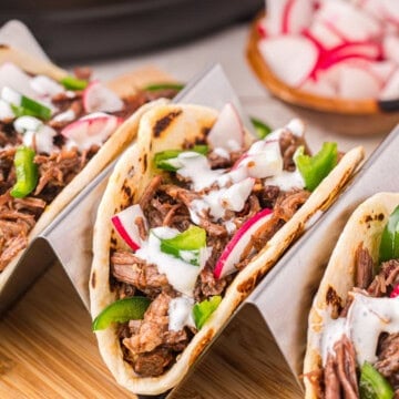 Barbacoa tacos in a taco holder on a wooden cutting board.