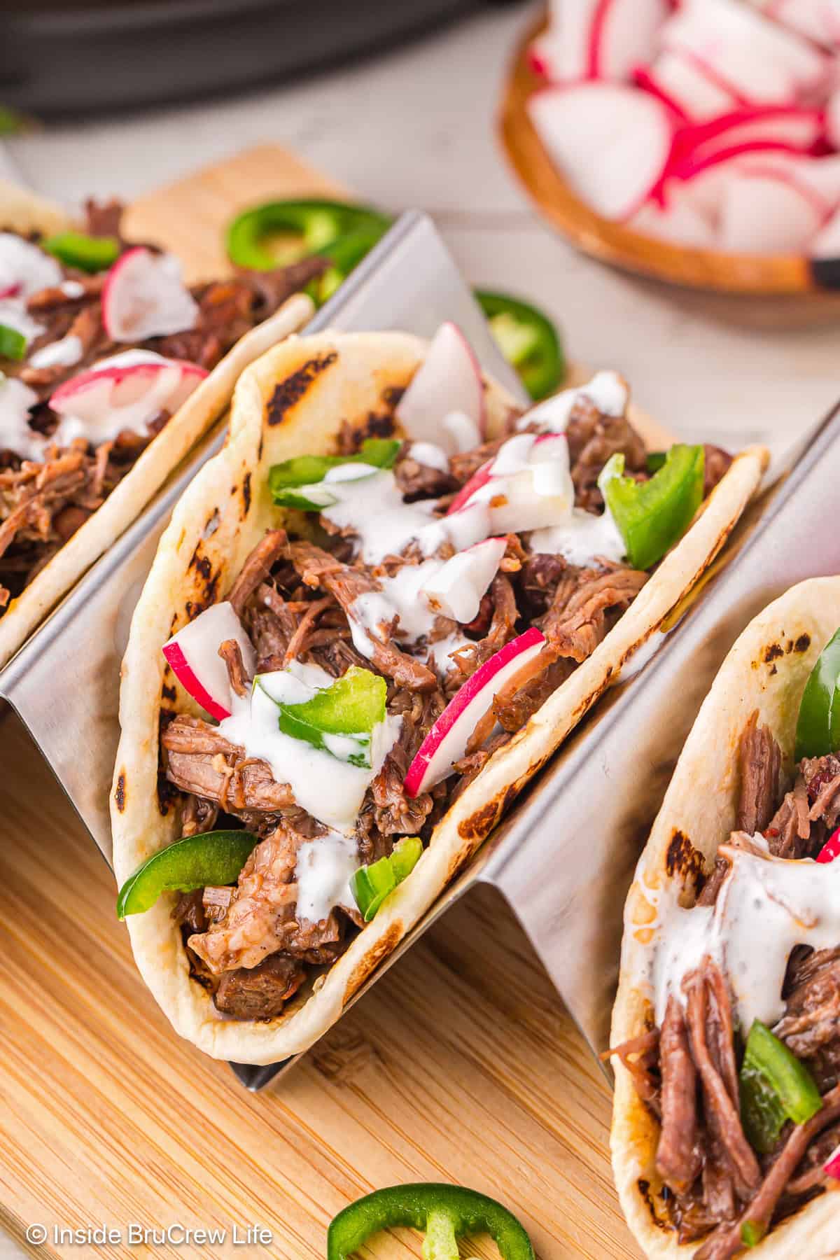 This easy barbacoa tacos recipe is made with juicy pulled beef in the Instant Pot! Delicious tucked into warm tortillas with toppings. Barbacoa tacos in a taco holder on a wooden cutting board.