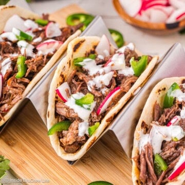 Barbacoa tacos in a taco holder on a wooden cutting board.