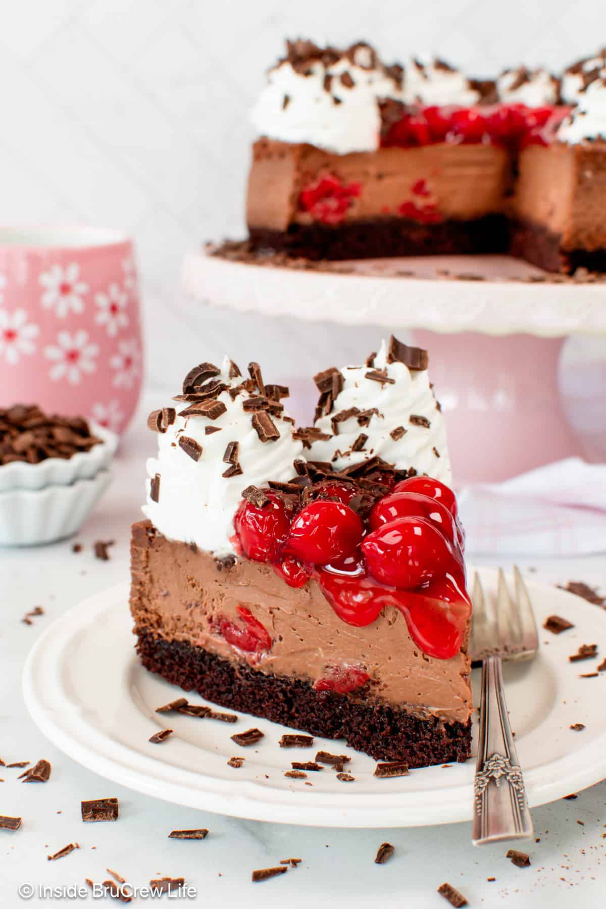 A slice of black forest cheesecake on a plate, showing the chocolate cheesecake and cake layers, topped with whipped cream and more cherries.