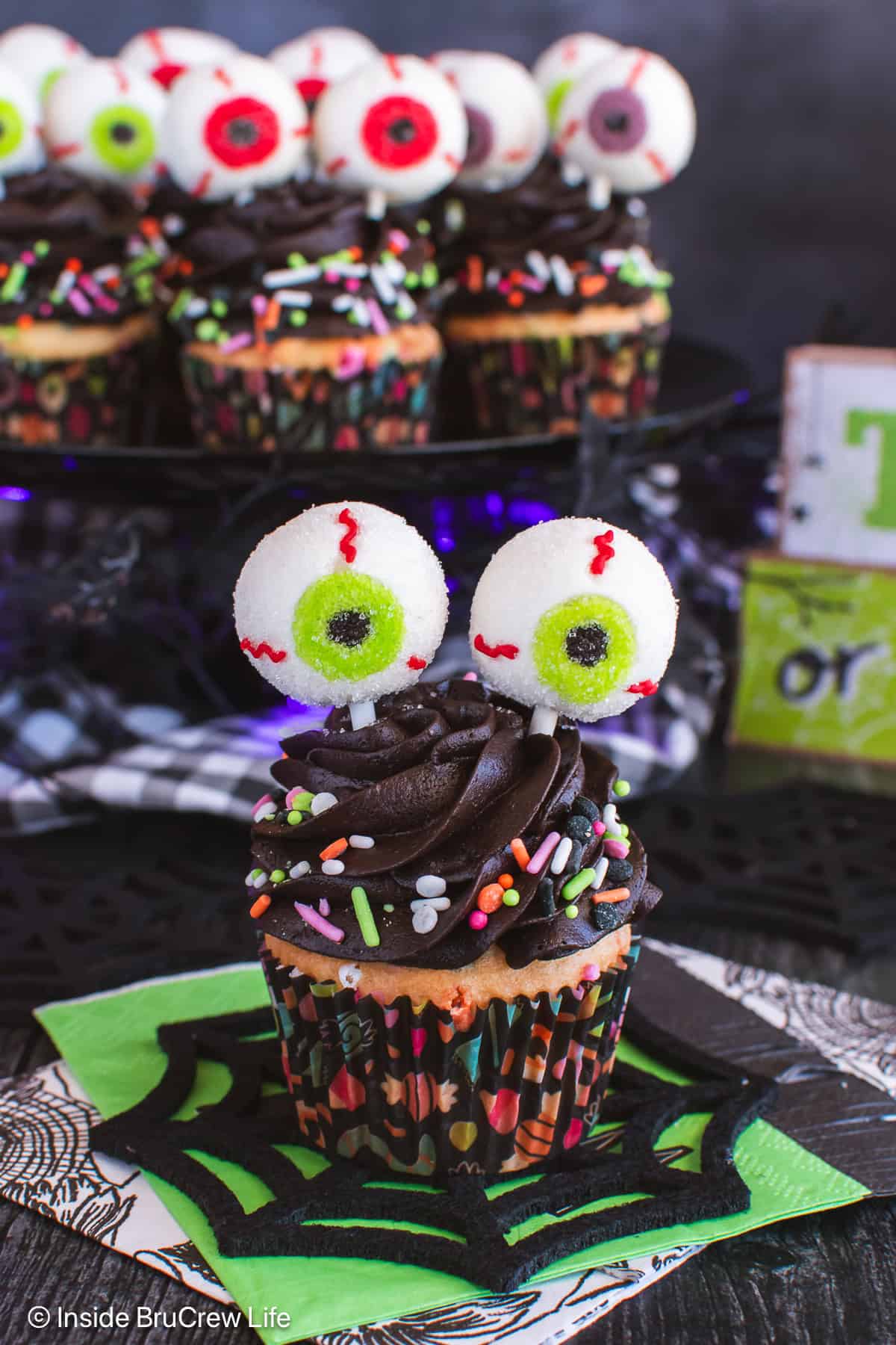An eyeball cupcake on a black spiderweb doily with a tray of cupcakes behind it.