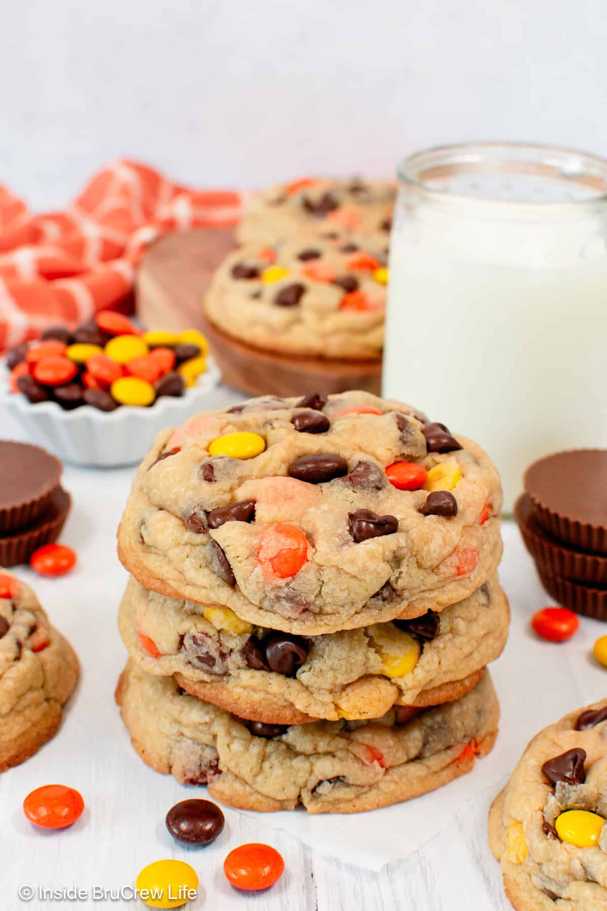 Chewy Reese's chocolate chip cookies stacked on a countertop with a glass of milk and more cookies in the background.