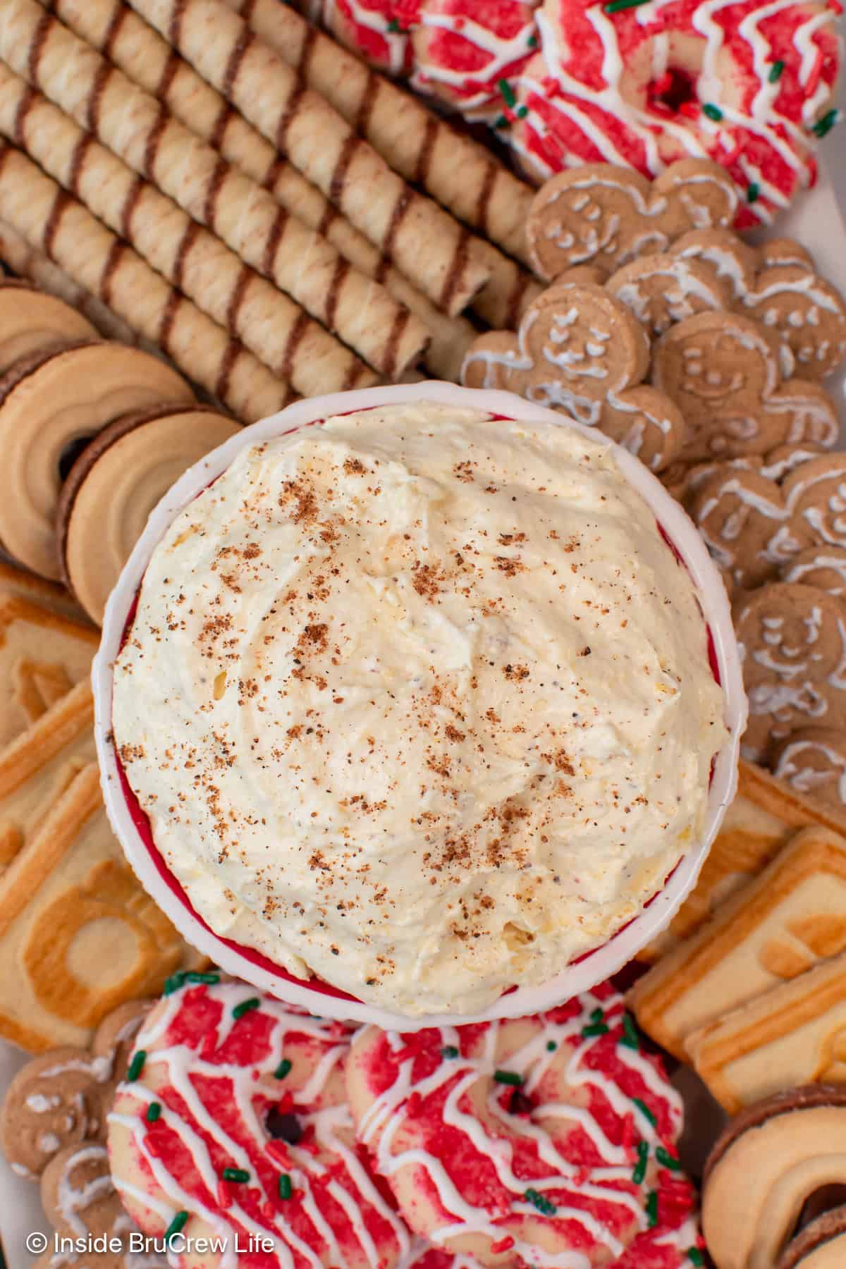 Light and fluffy eggnog dip is the easiest Christmas dessert made with instant pudding, eggnog, and whipped cream. It's ready in 10 minutes! Overhead view of a bowl of creamy eggnog dip garnished with nutmeg, surrounded by a platter of Christmas cookies.