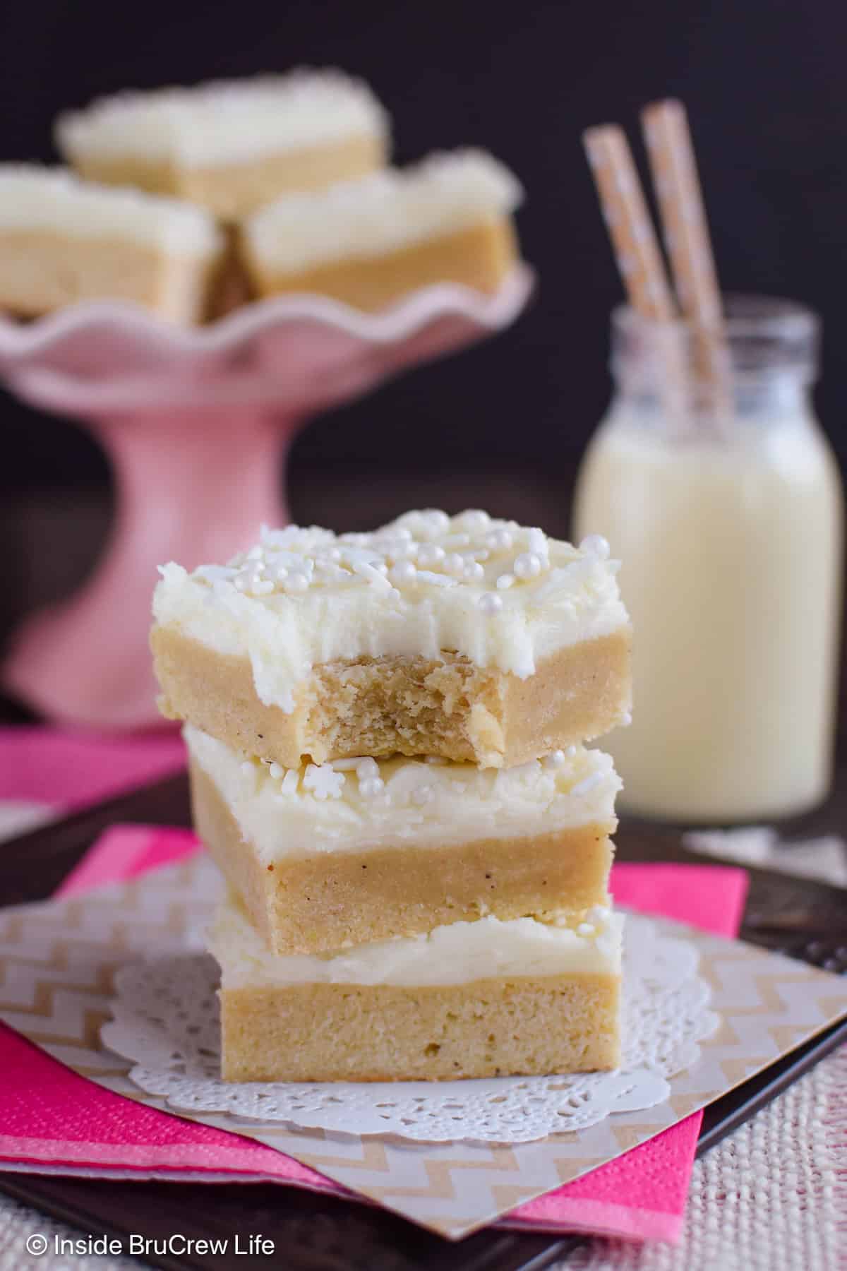 Three sugar cookie bars with eggnog frosting on a paper with a bite out of the top one.