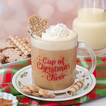 An eggnog latte topped with a swirl of whipped cream and a mini gingerbread man in a festive mug, set on top of a red and green plaid cloth.