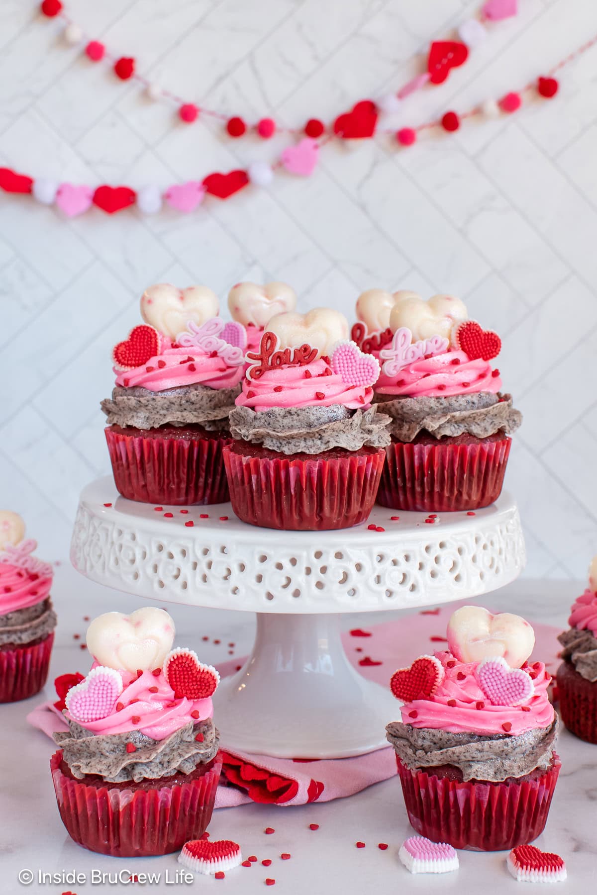 A white cake plate with red velvet Valentine's day cupcakes on it and around it.