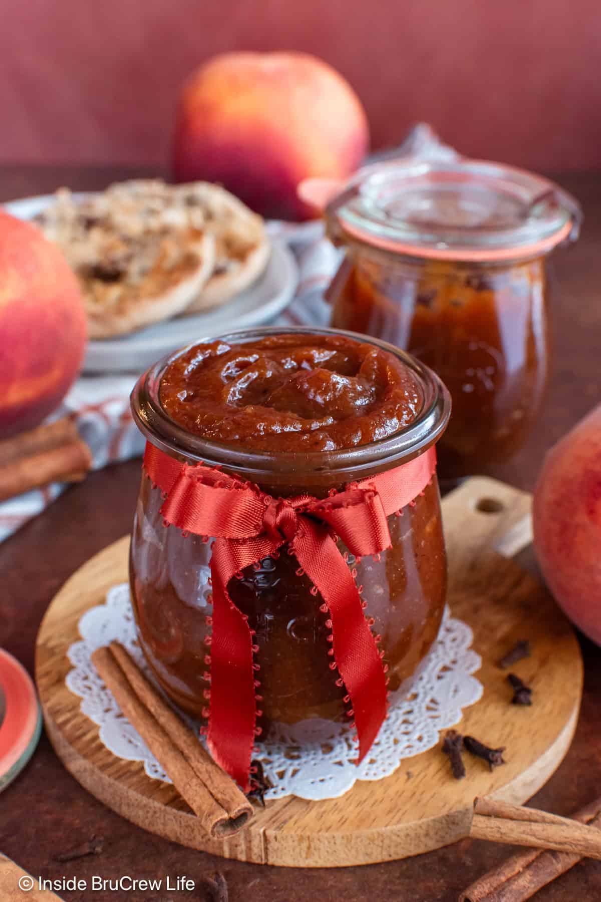 Two jars of peach butter with peaches and muffins behind it.